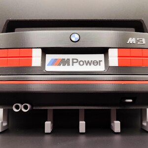 May include: A black model car rear end with red, white, and yellow taillights. The car has the text "M3" and a BMW logo. The license plate reads "M Power". The model has two exhaust pipes and a spoiler.