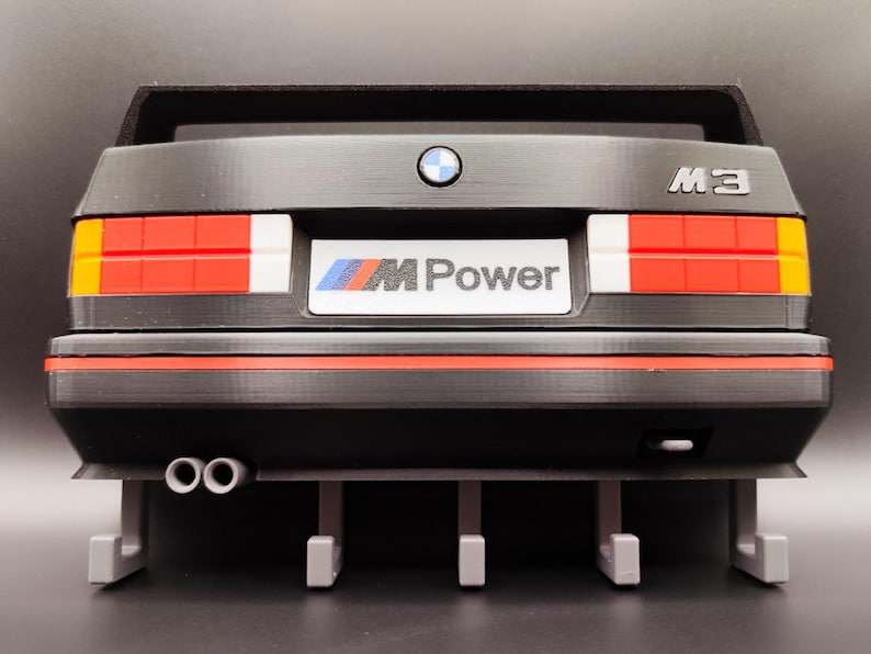 May include: A black model car rear end with red, white, and yellow taillights. The car has the text "M3" and a BMW logo. The license plate reads "M Power". The model has two exhaust pipes and a spoiler.
