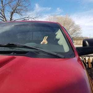 Puede incluir: Primer plano de una camioneta roja con un espejo lateral negro. Una mano dentro de la camioneta muestra el signo de la paz. El cielo es azul con nubes y un &aacute;rbol al fondo.