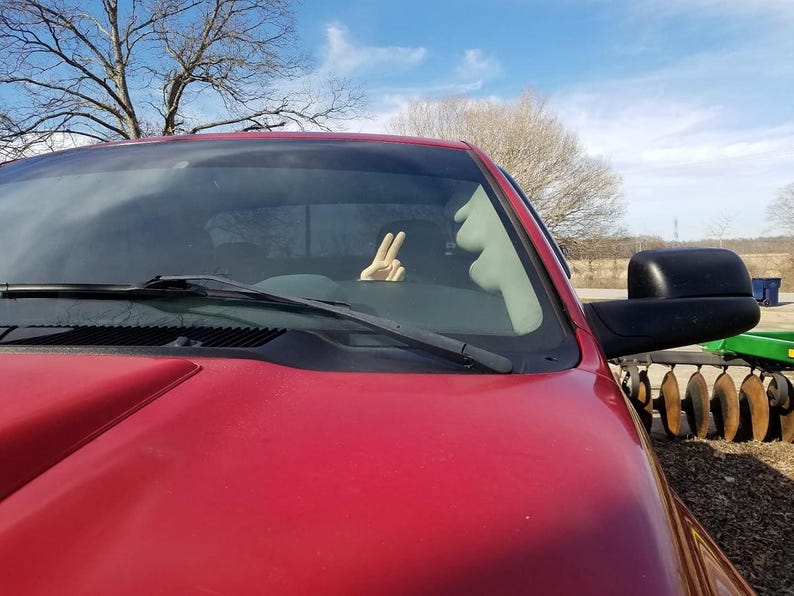 Puede incluir: Primer plano de una camioneta roja con un espejo lateral negro. Una mano dentro de la camioneta muestra el signo de la paz. El cielo es azul con nubes y un &aacute;rbol al fondo.