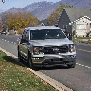 Puede incluir: Una camioneta plateada estacionada al costado de una carretera. La camioneta tiene una parrilla y faros negros. El fondo incluye casas, &aacute;rboles y monta&ntilde;as bajo un cielo azul.