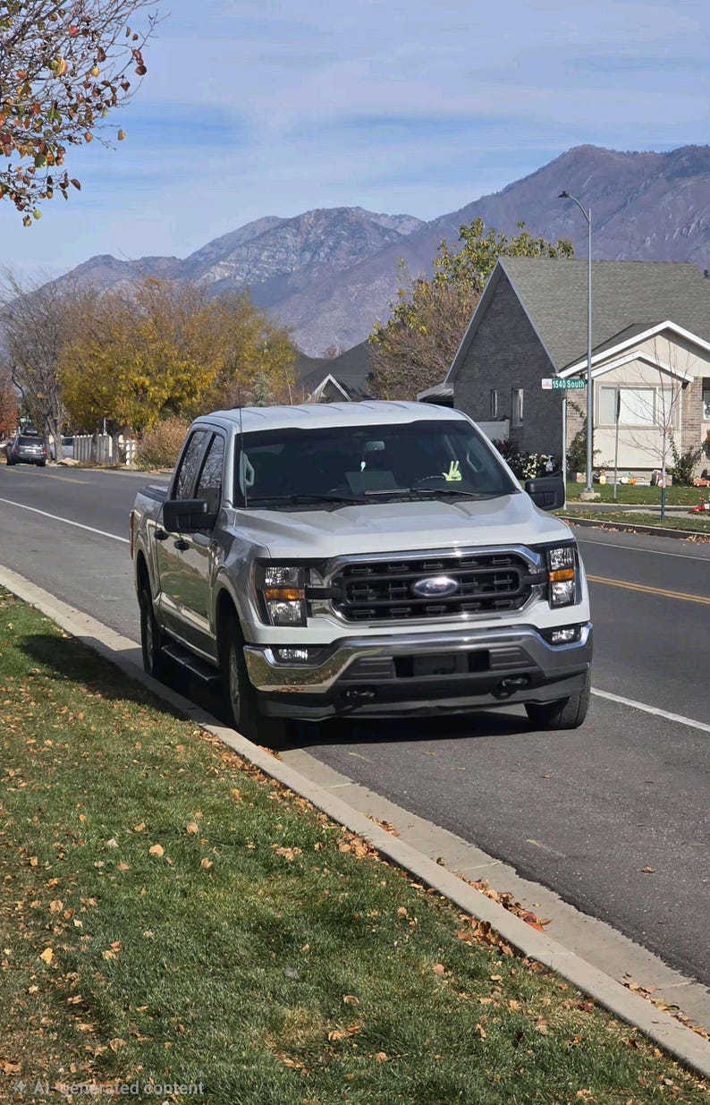 Puede incluir: Una camioneta plateada estacionada al costado de una carretera. La camioneta tiene una parrilla y faros negros. El fondo incluye casas, &aacute;rboles y monta&ntilde;as bajo un cielo azul.