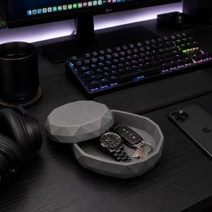 Puede incluir: Una configuraci&oacute;n de escritorio negra con teclado, rat&oacute;n y monitor. Una bandeja de baratijas gris geom&eacute;trica contiene un reloj, llaves y una llave de coche. Un iPhone negro y auriculares tambi&eacute;n est&aacute;n en el escritorio. Una taza negra est&aacute; sobre un posavasos.