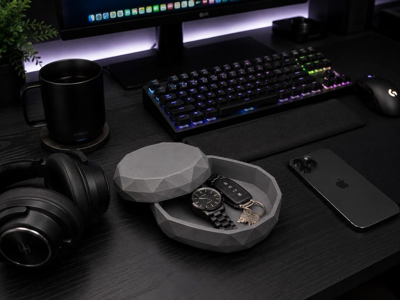Puede incluir: Una configuraci&oacute;n de escritorio negra con teclado, rat&oacute;n y monitor. Una bandeja de baratijas gris geom&eacute;trica contiene un reloj, llaves y una llave de coche. Un iPhone negro y auriculares tambi&eacute;n est&aacute;n en el escritorio. Una taza negra est&aacute; sobre un posavasos.