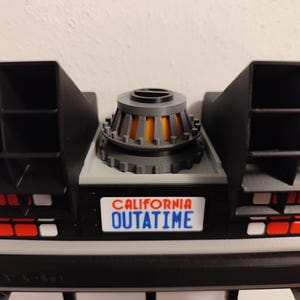 Pode incluir: Um modelo impresso em 3D da se&ccedil;&atilde;o traseira da m&aacute;quina do tempo DeLorean. O modelo apresenta aberturas pretas, uma base cinza e um reator central com detalhes em laranja. A placa diz "CALIFORNIA OUTATIME".