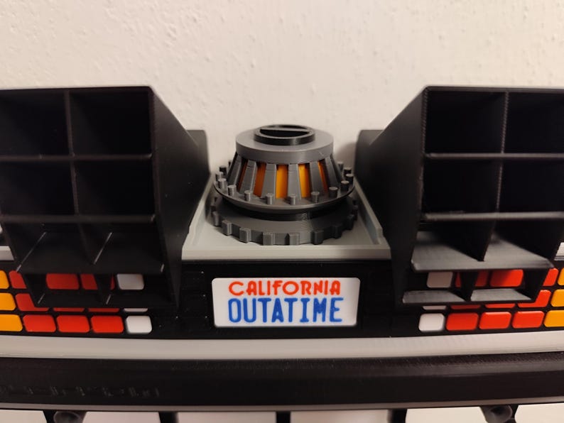 Pode incluir: Um modelo impresso em 3D da se&ccedil;&atilde;o traseira da m&aacute;quina do tempo DeLorean. O modelo apresenta aberturas pretas, uma base cinza e um reator central com detalhes em laranja. A placa diz "CALIFORNIA OUTATIME".