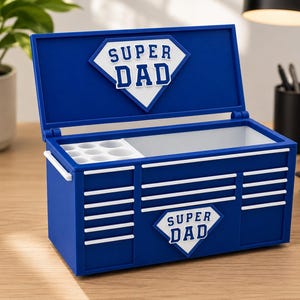 May include: A blue toolbox-shaped container with the words "SUPER DAD" in white. The container has a hinged lid and white accents. Inside, there are compartments for storage. The toolbox is on a wooden surface.