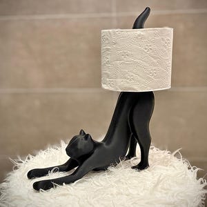Pode incluir: Um porta-papel higi&ecirc;nico em forma de gato preto. O gato est&aacute; em uma pose de alongamento, sustentando um rolo de papel higi&ecirc;nico branco estampado. O suporte est&aacute; em uma superf&iacute;cie branca e fofa.