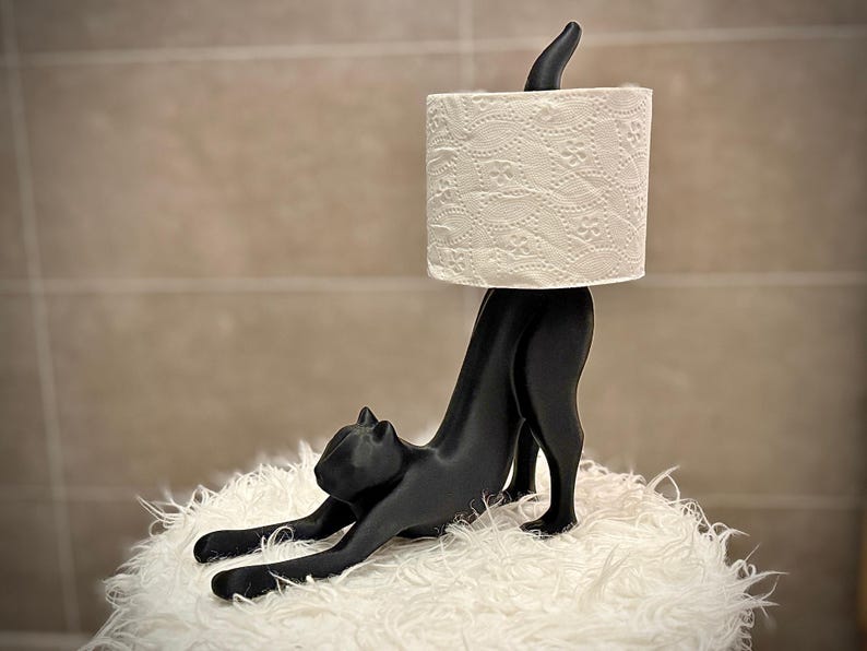 Pode incluir: Um porta-papel higi&ecirc;nico em forma de gato preto. O gato est&aacute; em uma pose de alongamento, sustentando um rolo de papel higi&ecirc;nico branco estampado. O suporte est&aacute; em uma superf&iacute;cie branca e fofa.