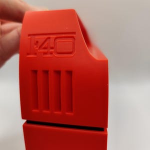 May include: Bright red plastic object with the number "1:40" embossed on the top. The object has a series of vertical lines on the side and a black base. The object is held by a hand.