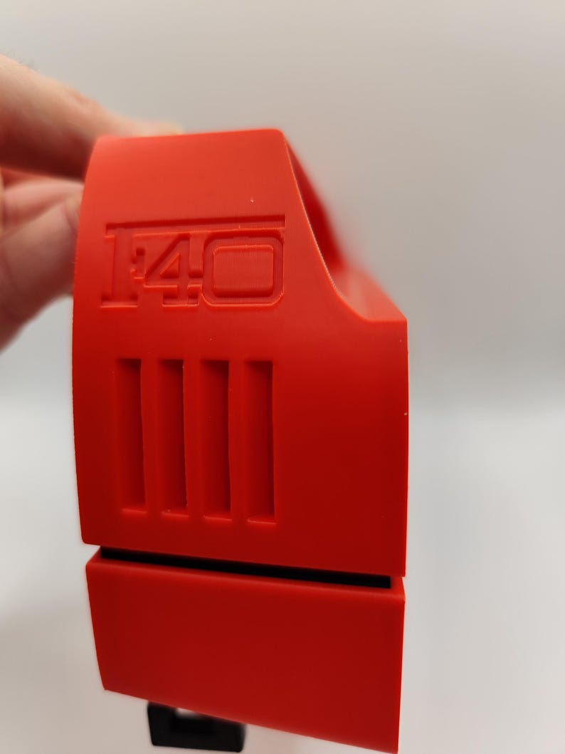 May include: Bright red plastic object with the number "1:40" embossed on the top. The object has a series of vertical lines on the side and a black base. The object is held by a hand.
