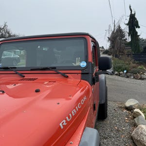 Puede incluir: Un SUV Jeep Rubicon rojo estacionado en un camino de grava. El veh&iacute;culo tiene un techo negro y guardabarros grises. La palabra "RUBICON" est&aacute; impresa en plata en el cap&oacute;.