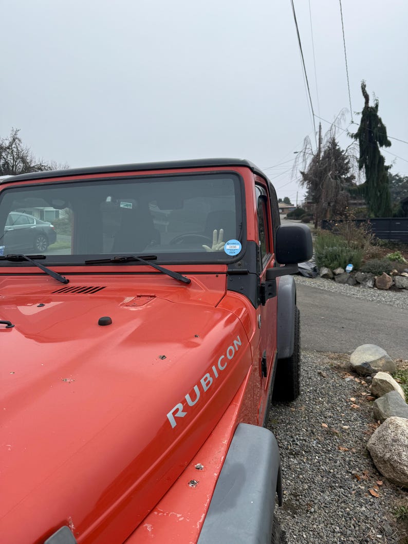 Puede incluir: Un SUV Jeep Rubicon rojo estacionado en un camino de grava. El veh&iacute;culo tiene un techo negro y guardabarros grises. La palabra "RUBICON" est&aacute; impresa en plata en el cap&oacute;.