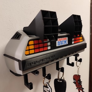 Pode incluir: Um porta-chaves DeLorean cinza e preto montado na parede. O porta-chaves tem uma placa de matr&iacute;cula que diz "OUTATIME" e apresenta v&aacute;rios ganchos para chaves. As luzes traseiras s&atilde;o vermelhas, laranja e amarelas.