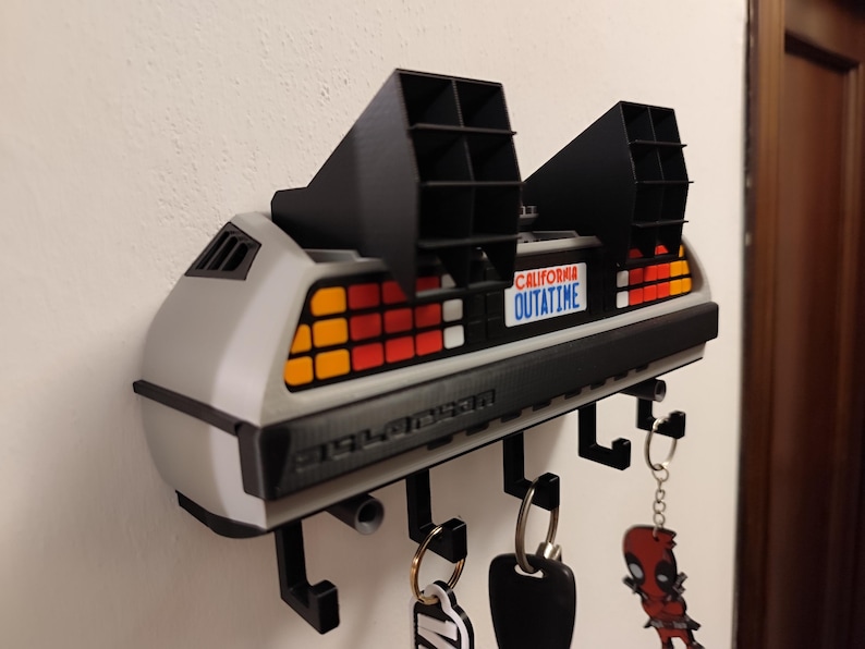 Pode incluir: Um porta-chaves DeLorean cinza e preto montado na parede. O porta-chaves tem uma placa de matr&iacute;cula que diz "OUTATIME" e apresenta v&aacute;rios ganchos para chaves. As luzes traseiras s&atilde;o vermelhas, laranja e amarelas.