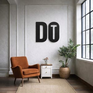Op de afbeelding: Zwarte metalen wandkunst met het woord "DO" in een modern lettertype. De letters zijn gemonteerd op een witte muur met textuur. Een oranje fauteuil, een kleine witte nachtkast en een plant in een pot staan op de voorgrond.