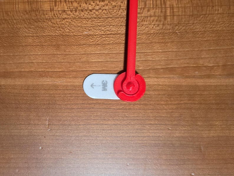 May include: A red 3D-printed object with a circular design and a stem, attached to a white, oval-shaped piece with the "3M" logo and an arrow. The object is set against a wooden surface.