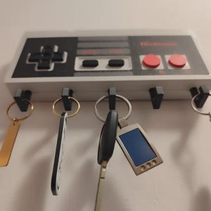 May include: A key holder shaped like a Nintendo controller, with a grey and black design. It has black buttons, red circular buttons, and a directional pad. Keys hang from the hooks below, including a gold rectangular key and a key with a blue screen.