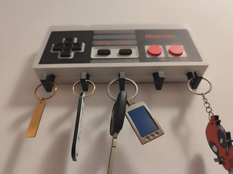 May include: A key holder shaped like a Nintendo controller, with a grey and black design. It has black buttons, red circular buttons, and a directional pad. Keys hang from the hooks below, including a gold rectangular key and a key with a blue screen.