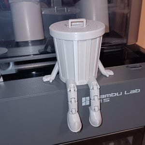Puede incluir: Un cubo de basura gris impreso en 3D con piernas y brazos articulados. El cubo de basura est&aacute; sentado en una superficie gris con el texto "Bambu Lab" impreso en &eacute;l.