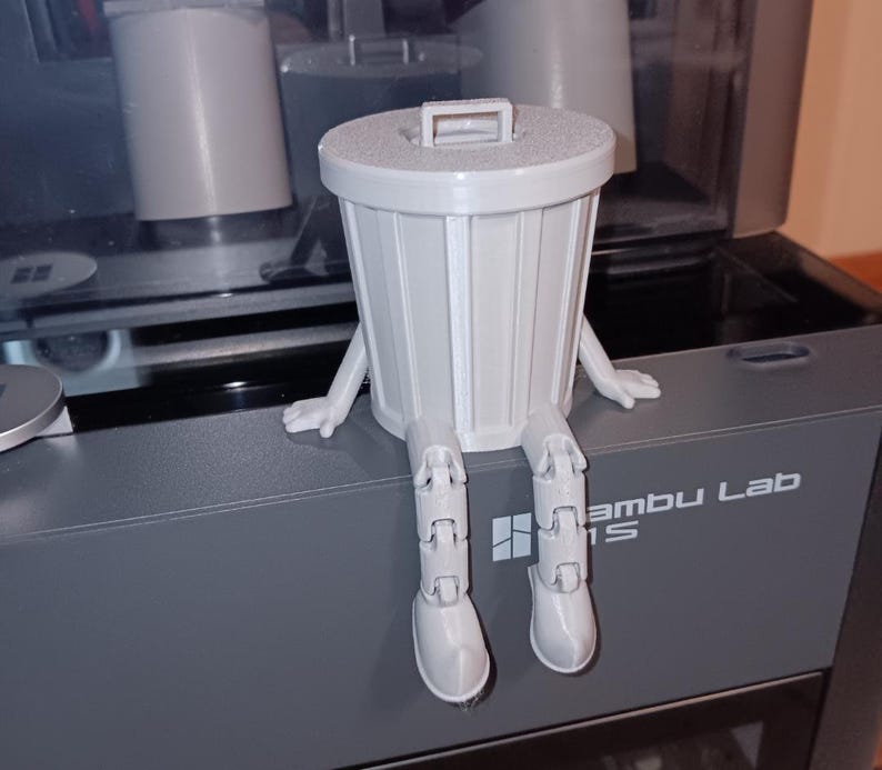 Puede incluir: Un cubo de basura gris impreso en 3D con piernas y brazos articulados. El cubo de basura est&aacute; sentado en una superficie gris con el texto "Bambu Lab" impreso en &eacute;l.