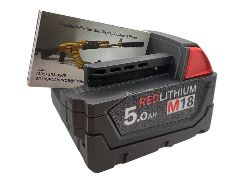 May include: A black and red Milwaukee M18 RedLithium 5.0Ah battery. The battery is on a black surface with a business card in the background.
