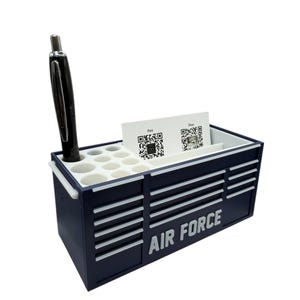 May include: Navy blue desk organizer shaped like a toolbox, with the words "AIR FORCE" in white. It has compartments for pens, business cards, and other small items. A black pen is in one of the pen holders.
