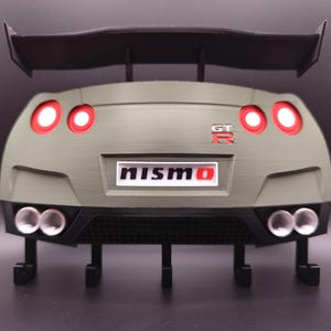 Może przedstawiać: Model 3D tylnej części samochodu, z czarnym spojlerem, czerwonymi światłami tylnymi i napisem "NISMO". Model jest oliwkowozielony z czarnymi akcentami i ma cztery rury wydechowe.