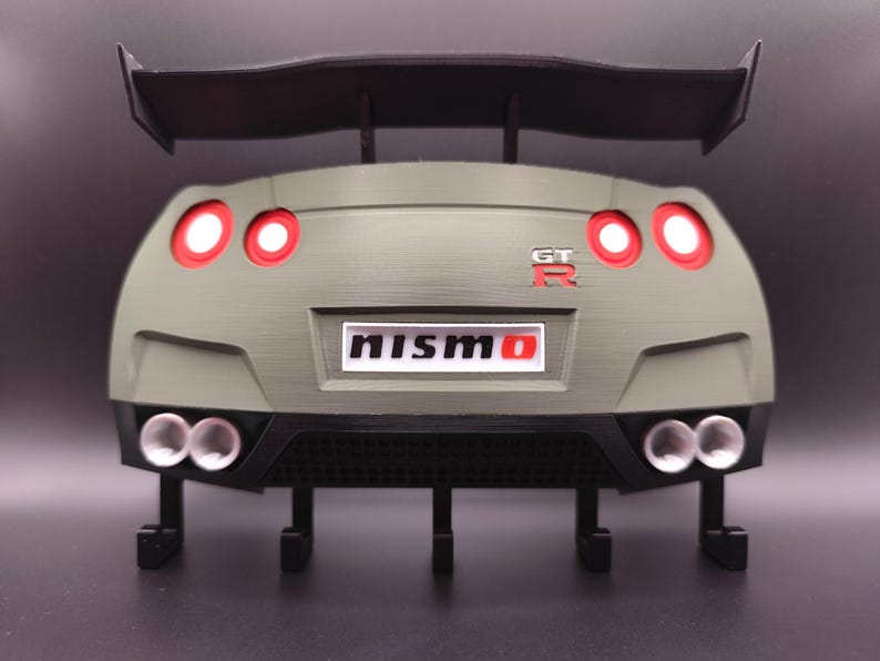 Może przedstawiać: Model 3D tylnej części samochodu, z czarnym spojlerem, czerwonymi światłami tylnymi i napisem "NISMO". Model jest oliwkowozielony z czarnymi akcentami i ma cztery rury wydechowe.