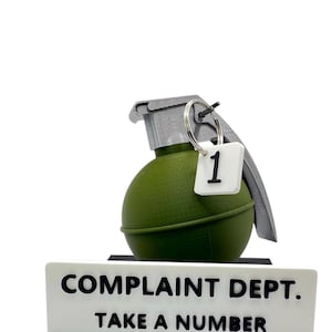 May include: A novelty desk sign with a green grenade and a tag with the number 1. The sign reads "COMPLAINT DEPT. TAKE A NUMBER" in black letters on a white background. The grenade has a silver pull pin.