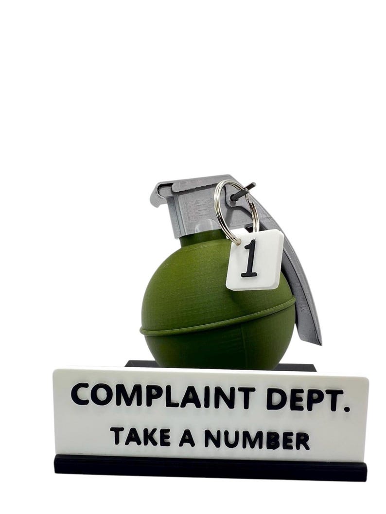 May include: A novelty desk sign with a green grenade and a tag with the number 1. The sign reads "COMPLAINT DEPT. TAKE A NUMBER" in black letters on a white background. The grenade has a silver pull pin.