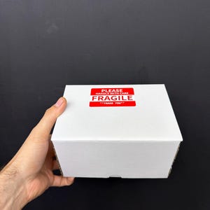 May include: A white cardboard box with a red "FRAGILE" sticker. The sticker reads "PLEASE HANDLE WITH CARE" and "THANK YOU". The box is held by a hand against a black background.