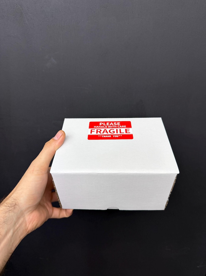 May include: A white cardboard box with a red "FRAGILE" sticker. The sticker reads "PLEASE HANDLE WITH CARE" and "THANK YOU". The box is held by a hand against a black background.