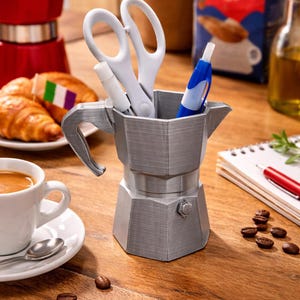 May include: A silver-colored desk organizer shaped like a stovetop espresso maker, holding white scissors, a white marker, and a blue pen. The organizer sits on a wooden surface, with coffee beans and a cup of espresso nearby.