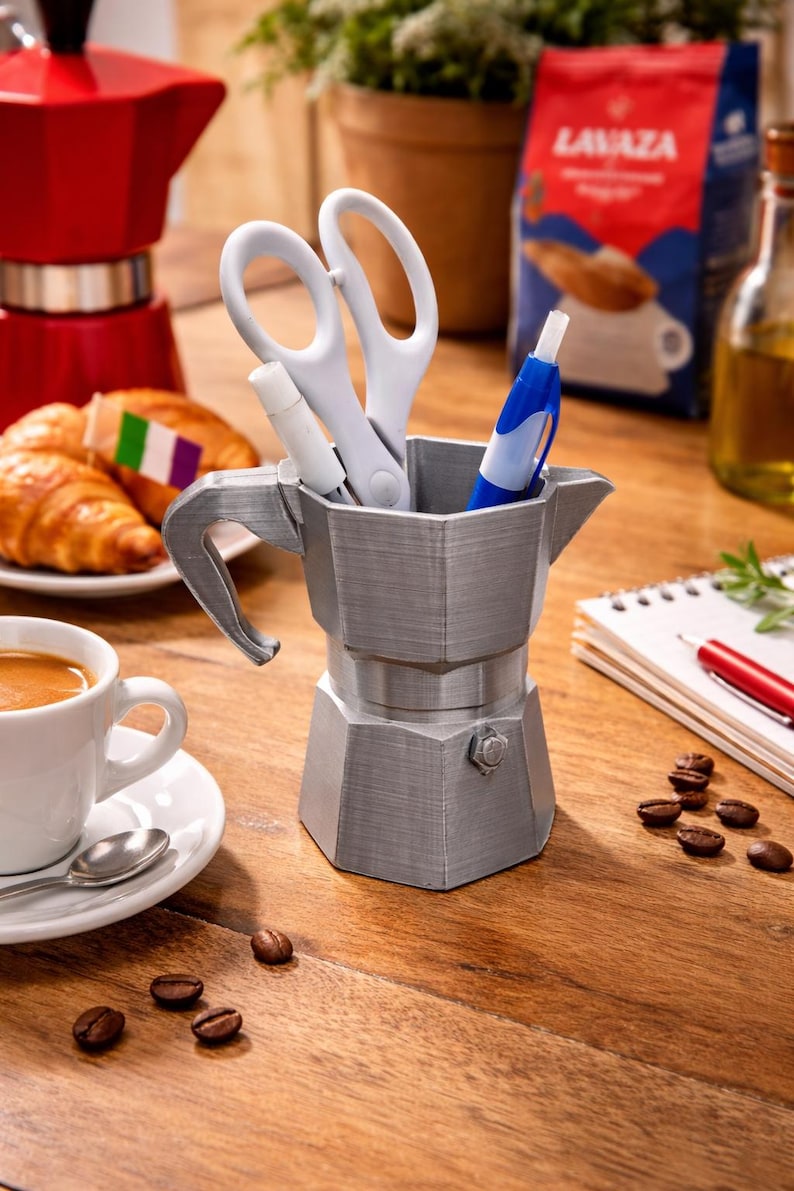 May include: A silver-colored desk organizer shaped like a stovetop espresso maker, holding white scissors, a white marker, and a blue pen. The organizer sits on a wooden surface, with coffee beans and a cup of espresso nearby.