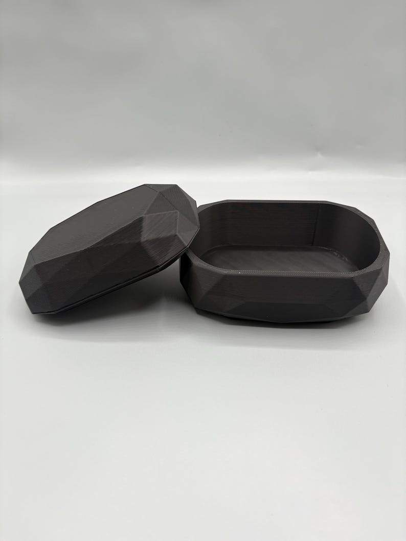 Puede incluir: Una caja de baratijas de color gris oscuro con forma geom&eacute;trica y tapa. La caja est&aacute; abierta, revelando el interior. La tapa se coloca a un lado. La caja y la tapa tienen un dise&ntilde;o facetado, lo que les da una apariencia moderna y angular. La caja es probablemente de pl&aacute;stico.