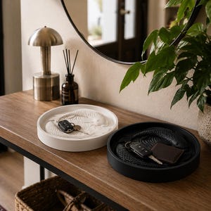Puede incluir: Dos bandejas decorativas sobre una mesa de madera. Una es blanca con un dise&ntilde;o texturizado y llaves, la otra es negra con un dise&ntilde;o texturizado, llaves y una peque&ntilde;a cartera marr&oacute;n. Una l&aacute;mpara, un difusor y una planta est&aacute;n en el fondo.