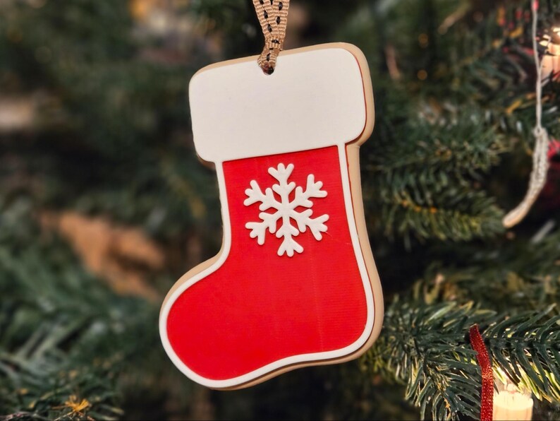 May include: A Christmas stocking ornament with a red body, white cuff, and a white snowflake design. The ornament is made of wood and has a brown ribbon for hanging. The background is a blurred Christmas tree.