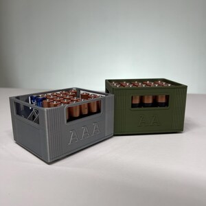 May include: Two battery storage containers, one grey and one olive green, each filled with batteries. The grey container is labelled "AAA", and the green container is labelled "AA". The batteries are arranged neatly inside the containers.