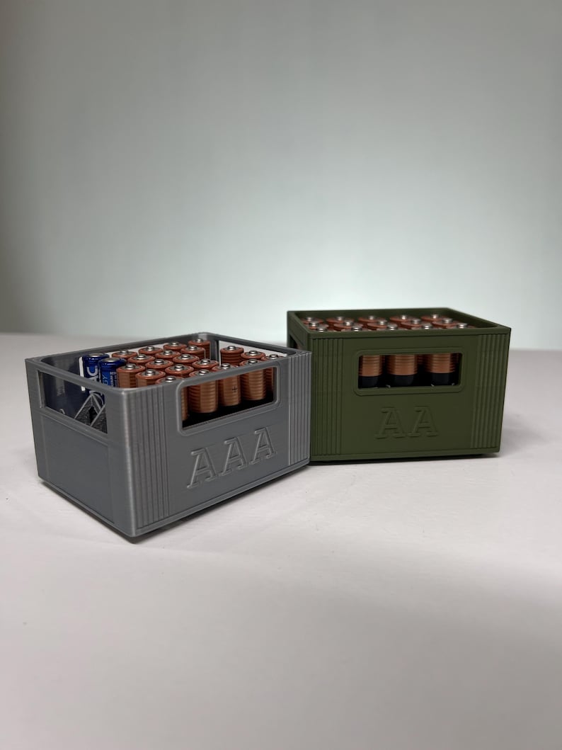 May include: Two battery storage containers, one grey and one olive green, each filled with batteries. The grey container is labelled "AAA", and the green container is labelled "AA". The batteries are arranged neatly inside the containers.