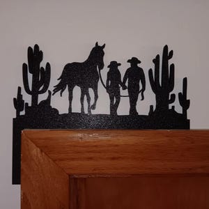 May include: Black metal silhouette of two people walking a horse with cacti in the background. The silhouette is mounted on a wooden door frame.