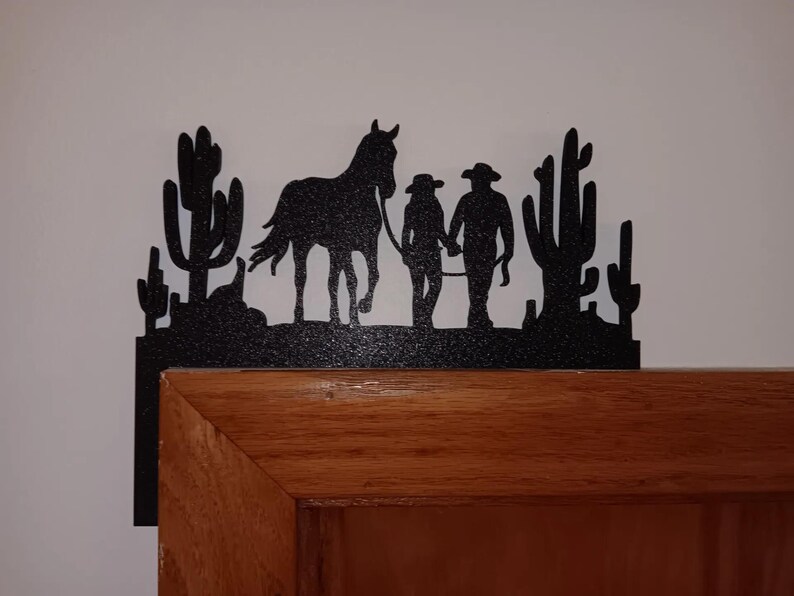 May include: Black metal silhouette of two people walking a horse with cacti in the background. The silhouette is mounted on a wooden door frame.