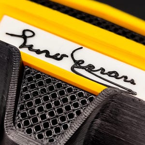 Puede incluir: Primer plano de un objeto negro y amarillo con el nombre "Liner Ferrari" en escritura negra sobre fondo blanco. El objeto tiene un patr&oacute;n texturizado en forma de rejilla y un borde amarillo.