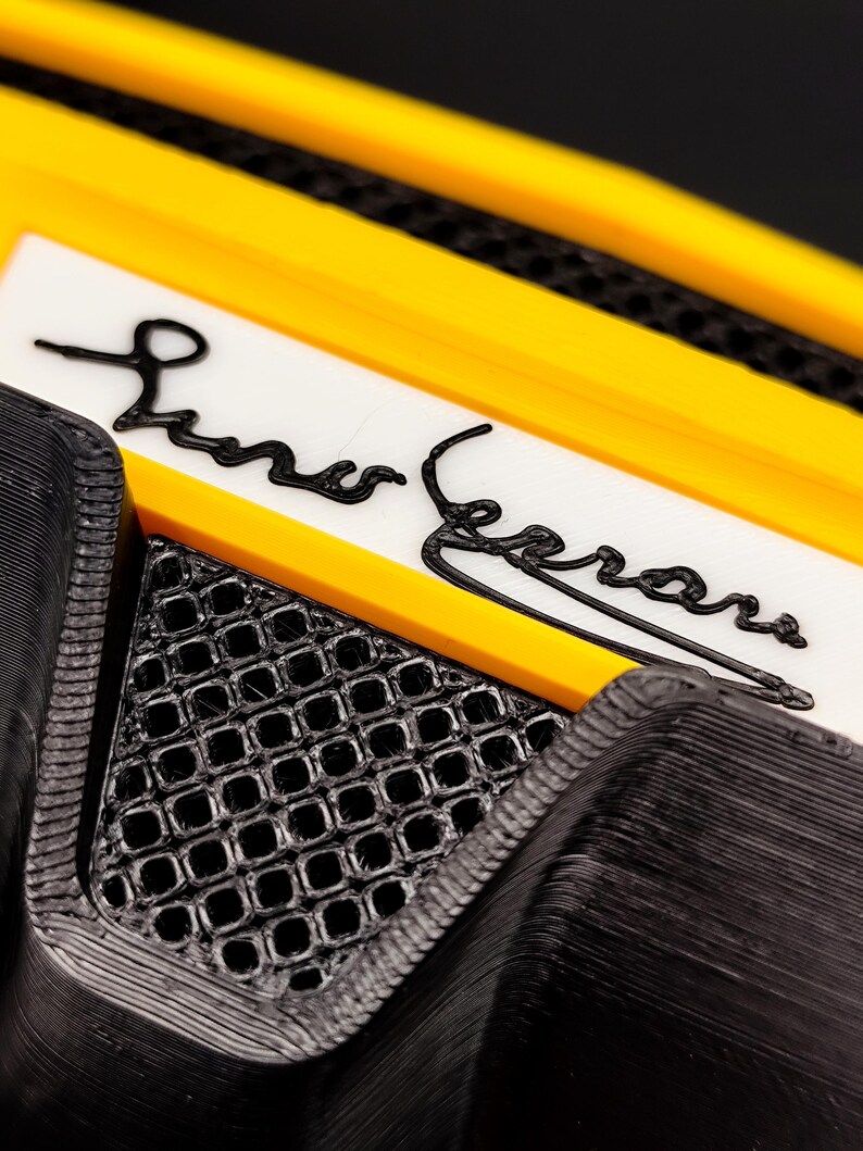 Puede incluir: Primer plano de un objeto negro y amarillo con el nombre "Liner Ferrari" en escritura negra sobre fondo blanco. El objeto tiene un patr&oacute;n texturizado en forma de rejilla y un borde amarillo.