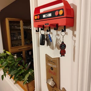 May include: A red key holder shaped like a car, mounted on a white door. The key holder has black hooks and holds keys and a keychain. The key holder has the number "140" on it. A wooden sign with a bottle opener is also visible.
