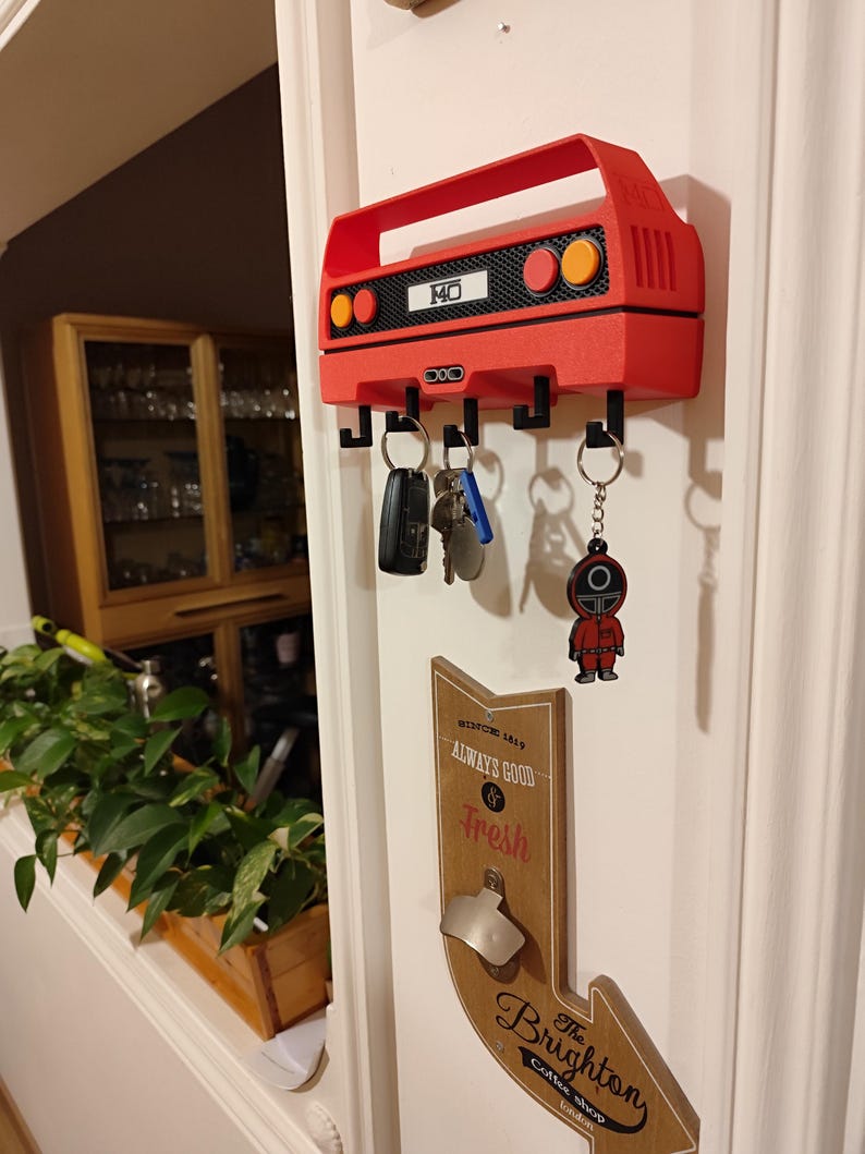May include: A red key holder shaped like a car, mounted on a white door. The key holder has black hooks and holds keys and a keychain. The key holder has the number "140" on it. A wooden sign with a bottle opener is also visible.