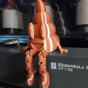 Puede incluir: Una figurita impresa en 3D de un emoji de caca sonriente con piernas articuladas. La figurita es de color marr&oacute;n y tiene un acabado brillante. La figurita est&aacute; sentada sobre una superficie negra con el texto "Bambu P1S" impreso en ella.