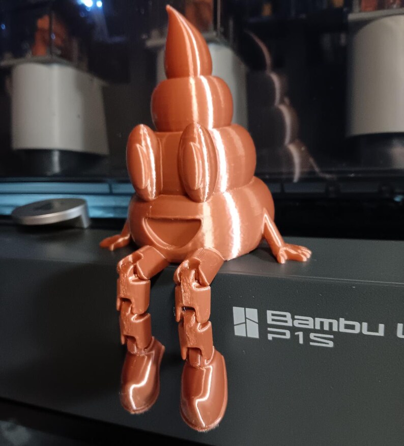 Puede incluir: Una figurita impresa en 3D de un emoji de caca sonriente con piernas articuladas. La figurita es de color marr&oacute;n y tiene un acabado brillante. La figurita est&aacute; sentada sobre una superficie negra con el texto "Bambu P1S" impreso en ella.