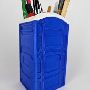 May include: A blue plastic desk organizer shaped like a portable toilet. The organizer has a white top and is filled with pens, pencils, and markers.