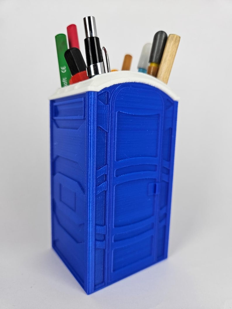 May include: A blue plastic desk organizer shaped like a portable toilet. The organizer has a white top and is filled with pens, pencils, and markers.